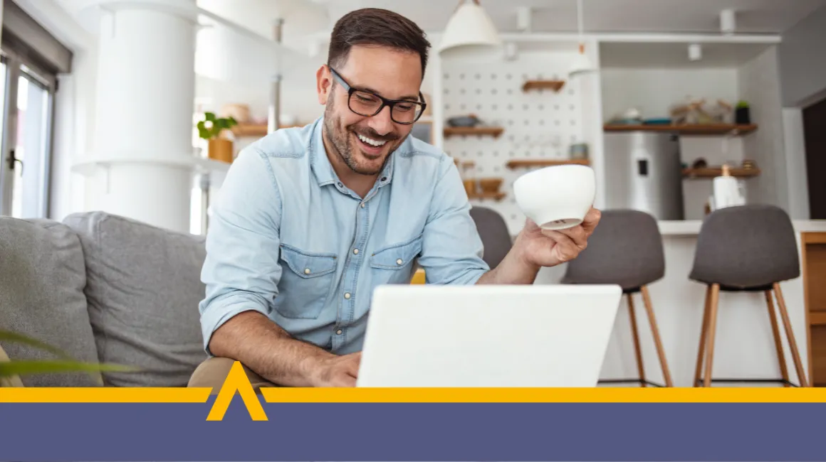 Uśmiechnięty mężczyzna korzystający z laptopa w domowym wnętrzu, trzymający filiżankę